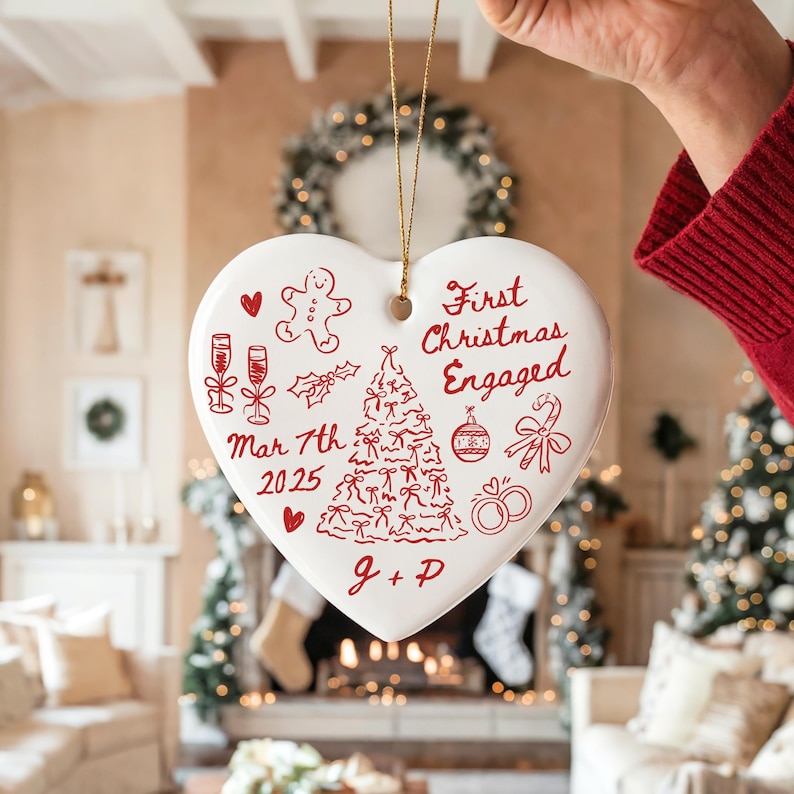 May include: A white heart-shaped Christmas ornament with red illustrations and text. The ornament features a Christmas tree, champagne glasses, and the words "First Christmas Engaged" and "Mar 7th 2025". The initials "J + P" are also included.