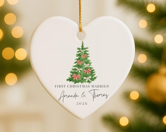 May include: Heart-shaped ceramic Christmas ornament with a watercolor Christmas tree design. The tree is decorated with pink bows. Text reads "First Christmas Married" with names and a year.