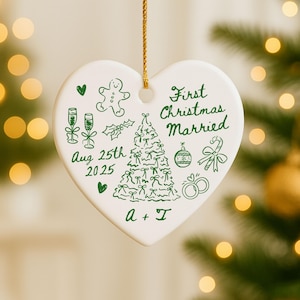 May include: A heart-shaped, white ceramic Christmas ornament with green illustrations and text. The ornament features a Christmas tree, champagne glasses, and the words "First Christmas Married" and "Aug 25th 2025". A gold string is attached for hanging.