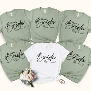 May include: A collection of six t-shirts, with the text "Team Bride" in a script font. Five shirts are a muted green, and one is white. A small bouquet of flowers and two gold rings are in the foreground.