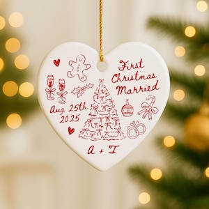 May include: A heart-shaped white ceramic Christmas ornament with red illustrations and text. The ornament features a Christmas tree, champagne glasses, and the words "First Christmas Married" and "Aug 25th 2025". A gold string is attached for hanging.