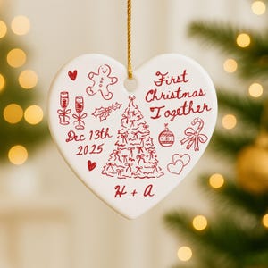May include: A heart-shaped Christmas ornament with red illustrations and text. The ornament features a Christmas tree, gingerbread man, champagne glasses, and the words "First Christmas Together" and "Dec 13th 2025". A gold string is attached for hanging.