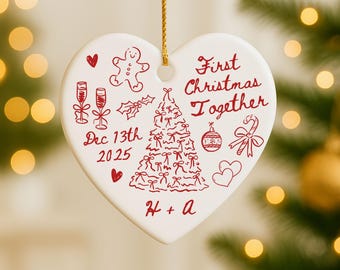 Könnte beinhalten: Ein herzförmiger Weihnachtsbaumschmuck mit roten Illustrationen und Text. Der Schmuck zeigt einen Weihnachtsbaum, einen Lebkuchenmann, Sektgläser und die Worte "First Christmas Together" und "Dec 13th 2025". Ein goldener Faden ist zum Aufhängen befestigt.