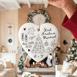 May include: A white heart-shaped Christmas ornament with black hand-drawn illustrations and text. The ornament says "First Christmas Married" and includes a date, a Christmas tree, and other festive drawings. It is held by a person.
