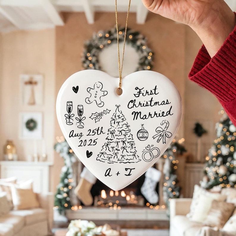 May include: A white heart-shaped Christmas ornament with black hand-drawn illustrations and text. The ornament says "First Christmas Married" and includes a date, a Christmas tree, and other festive drawings. It is held by a person.