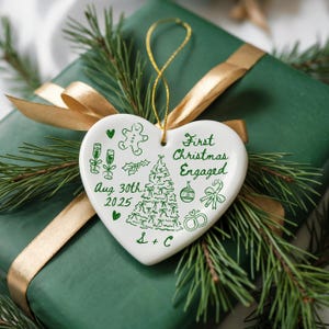 May include: A white heart-shaped Christmas ornament with green illustrations and text, including "First Christmas Engaged" and "Aug 30th 2025". The ornament hangs from a gold string and rests on a green wrapped gift with a gold ribbon and pine branches.