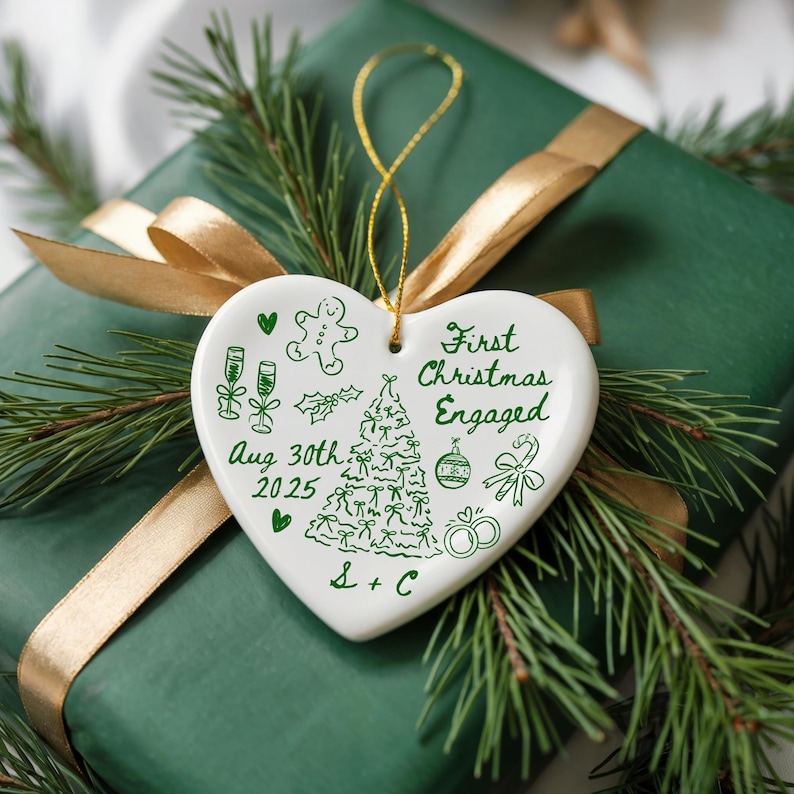 May include: A white heart-shaped Christmas ornament with green illustrations and text, including "First Christmas Engaged" and "Aug 30th 2025". The ornament hangs from a gold string and rests on a green wrapped gift with a gold ribbon and pine branches.