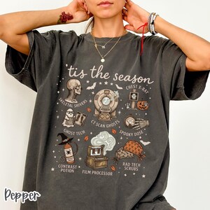 May include: A charcoal gray t-shirt featuring the text 'tis the season and various medical-themed Halloween illustrations including a skull, CT scanner, ghost, pumpkin, film processor, and scrub caps with labels like Skeletal Shivers and Ghost Tech.