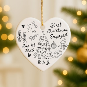 May include: White heart-shaped Christmas ornament with black hand-drawn illustrations and text. The ornament says "First Christmas Engaged" and includes a Christmas tree, champagne glasses, and the date "Aug 1st 2025".