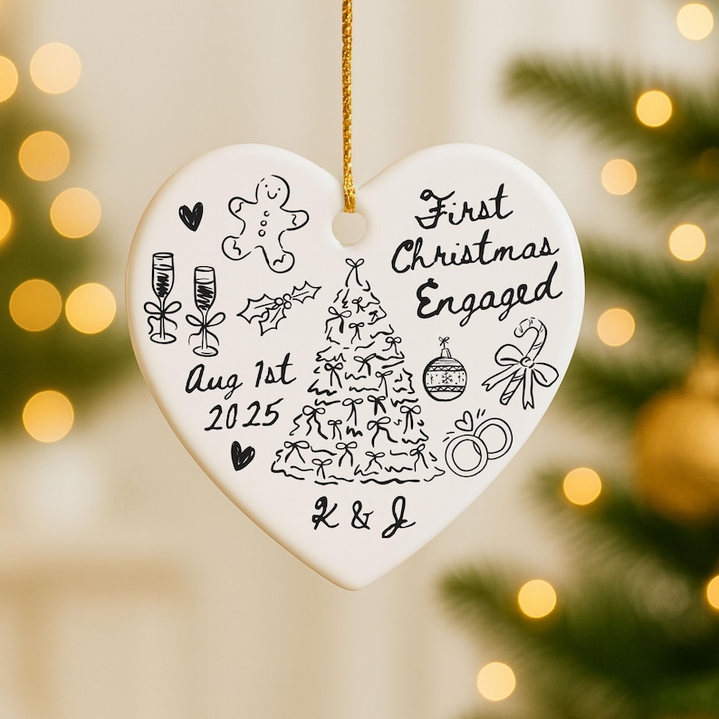May include: White heart-shaped Christmas ornament with black hand-drawn illustrations and text. The ornament says "First Christmas Engaged" and includes a Christmas tree, champagne glasses, and the date "Aug 1st 2025".
