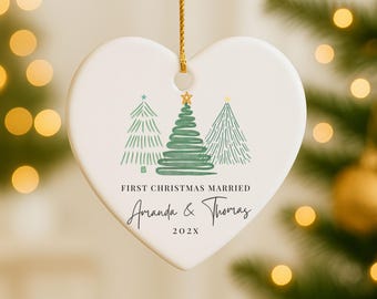 May include: Heart-shaped ceramic Christmas ornament with a gold string. The ornament features three green Christmas trees and the text "FIRST CHRISTMAS MARRIED" with the names "Amanda & Thomas" and the year "202X".