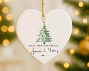 May include: Heart-shaped ceramic Christmas ornament with a watercolor Christmas tree design. The ornament has the text "FIRST CHRISTMAS MARRIED" and the names "Amanda & Thomas" with the year "202X". It hangs from a gold cord.