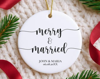 May include: White ceramic Christmas ornament with the words "merry & married" in black script. The names "John & Maria" and a date are printed below. A gold string is attached for hanging. Red ribbon and pine branches surround the ornament.