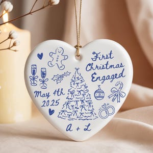 May include: White heart-shaped Christmas ornament with blue illustrations and text. The ornament says "First Christmas Engaged" and includes a Christmas tree, champagne glasses, and the date "May 4th 2025".