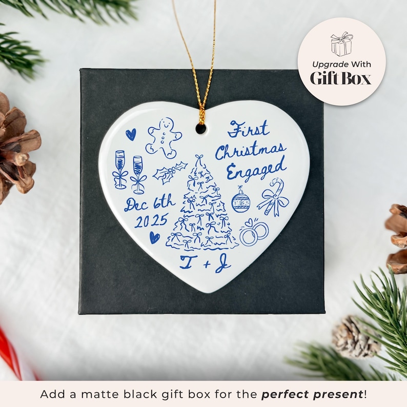 May include: A white heart-shaped Christmas ornament with blue illustrations and text. The ornament features a Christmas tree, champagne glasses, and the words "First Christmas Engaged" and "Dec 6th 2025". It rests on a black gift box.