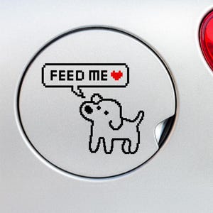 May include: A silver car fuel door with a black and white pixelated dog illustration. The dog has a speech bubble that says "FEED ME" with a red heart. The car's red tail light is visible in the background.