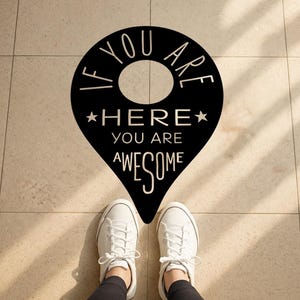 May include: A black floor decal in the shape of a location pin with the words "IF YOU ARE HERE YOU ARE AWESOME" in white. White sneakers are positioned in front of the decal on a tiled floor.