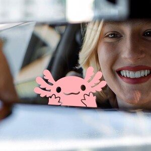 May include: A pink axolotl car sticker with a smiling face and frilly gills. The sticker is seen through a car window, with a smiling person in the background.