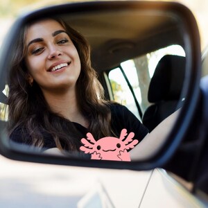May include: A pink axolotl sticker is seen in a car's wing mirror. The sticker features a smiling face and is placed near the mirror's base. The reflection shows a person smiling while driving.