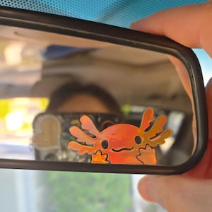 May include: A car's rearview mirror with an orange axolotl sticker. The sticker features a smiling face and detailed gills. The mirror is held by a hand, with a blurred interior visible in the background.