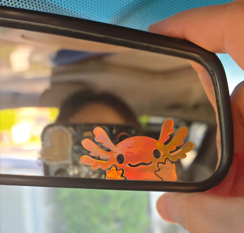 May include: A car's rearview mirror with an orange axolotl sticker. The sticker features a smiling face and detailed gills. The mirror is held by a hand, with a blurred interior visible in the background.