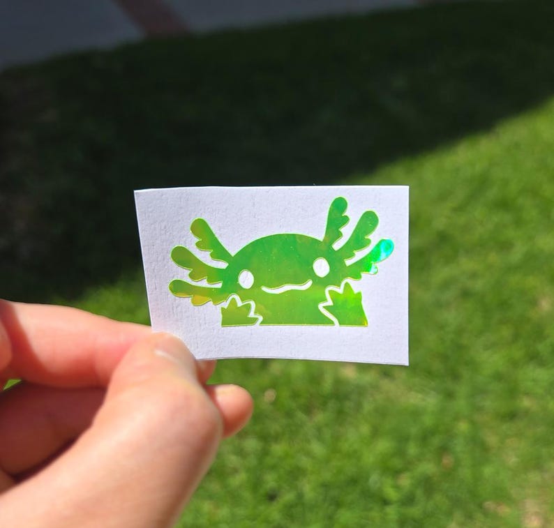 May include: A holographic sticker with a cartoon axolotl design. The axolotl is green with a smiling face and frilly gills. The sticker is held against a background of green grass.