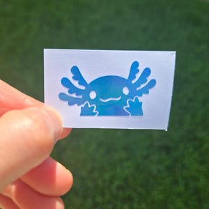 May include: A blue axolotl sticker with a holographic finish. The sticker depicts a cartoon axolotl with a smiling face and feathery gills. The sticker is held in a hand against a green background.