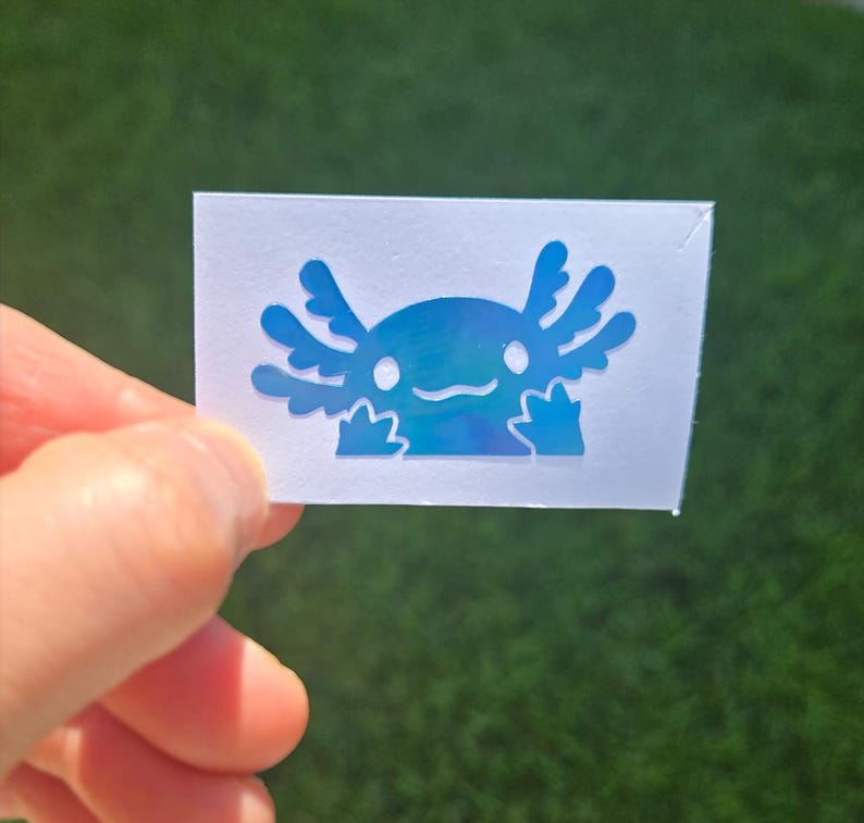 May include: A blue axolotl sticker with a holographic finish. The sticker depicts a cartoon axolotl with a smiling face and feathery gills. The sticker is held in a hand against a green background.