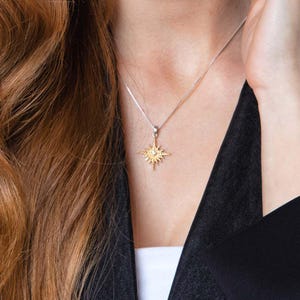 May include: A silver necklace with a gold-colored starburst pendant. The pendant has a small, clear gemstone in the center. The necklace is worn against a black top and white undershirt.