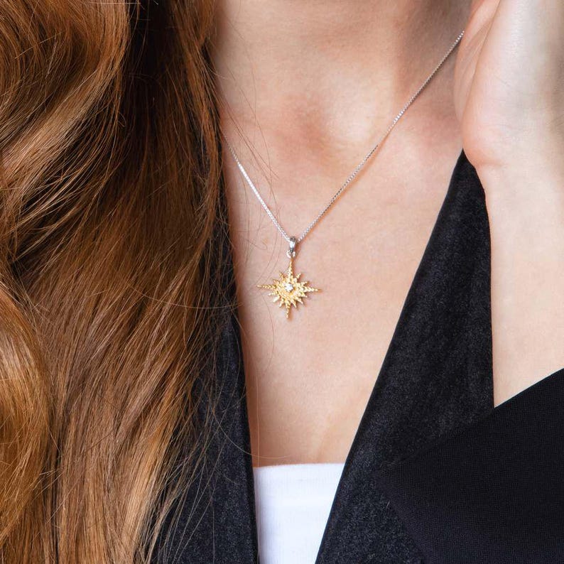 May include: A silver necklace with a gold-colored starburst pendant. The pendant has a small, clear gemstone in the center. The necklace is worn against a black top and white undershirt.