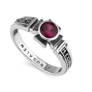 May include: A silver ring featuring a cross design and a central, round, deep red gemstone. The band is inscribed with "JERUSALEM". The ring also has text on the top and sides.