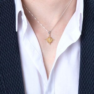 May include: A gold-colored pendant necklace with a starburst design, featuring a central gemstone. The necklace is worn over a white collared shirt and a dark blue blazer. The pendant hangs from a delicate silver chain.