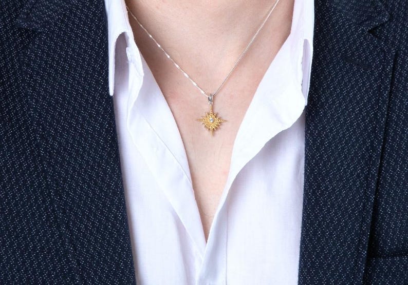 May include: A gold-colored pendant necklace with a starburst design, featuring a central gemstone. The necklace is worn over a white collared shirt and a dark blue blazer. The pendant hangs from a delicate silver chain.
