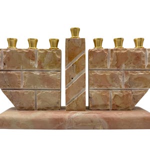 Może przedstawiać: Kamienna menora z prostokątną podstawą i centralną kolumną. Menora ma dziewięć świeczników ze złotymi nakładkami. Kamień to mieszanka odcieni beżu, różu i brązu, co nadaje mu naturalny, teksturowany wygląd.