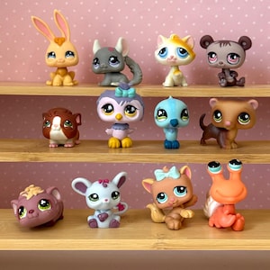 May include: A collection of eleven small plastic animal figurines with oversized eyes, including a rabbit, chinchilla, cat, bear, guinea pig, owl, bird, dog, mouse, and a hermit crab, arranged on three wooden shelves against a pink dotted background.