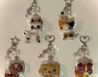 May include: Collection of keychains featuring miniature animal figurines, including dogs and cats. Each keychain has a silver-tone clasp and is adorned with decorative charms, bows, and beads in pastel colors. The figurines have large, expressive eyes.