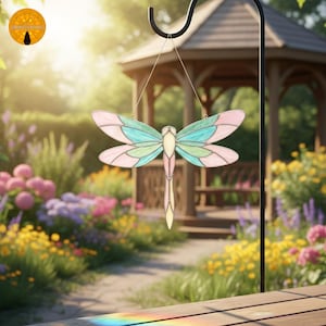 May include: A stained glass dragonfly with pink, teal, and green wings hangs from a black hook. The dragonfly is suspended in front of a blurred garden scene with a gazebo and colorful flowers. The dragonfly's wings are divided into sections.