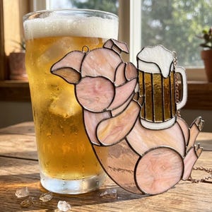 Puede incluir: Adorno de cerdo de vidriera sosteniendo una jarra de cerveza. El cerdo es rosa y blanco con una jarra de cerveza en sus brazos. Un vaso de cerveza está en el fondo. El adorno cuelga de una cadena.