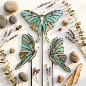 Può includere: Tre ornamenti in vetro colorato a forma di falena luna in tonalità verde pallido e turchese iridescente, ciascuno montato su una sottile asta di metallo scuro. Le ali presentano delicate linee di saldatura scure e pannelli di vetro traslucido.