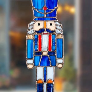 Könnte beinhalten: Ein Glasmalerei-Ornament in Form eines Nussknacker-Soldaten. Das Ornament weist Blautöne, Weiß, Rot und Gold auf. Der Soldat hat einen blauen Hut mit einem roten Edelstein, einen weißen Schnurrbart und eine rot-weiße Uniform. Eine goldene Kette ist zum Aufhängen befestigt.