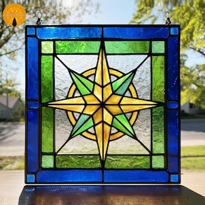 May include: Stained glass window panel featuring a star compass design. The panel has a square shape with a blue border, green and clear glass sections, and a central starburst in yellow and green. A decorative piece for home decor.