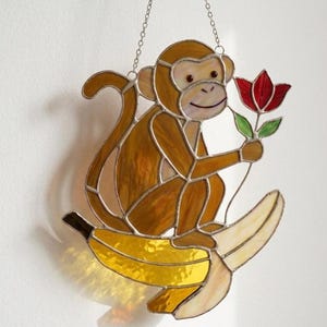 May include: Stained glass monkey holding a red flower, sitting on a banana. The monkey is brown with a light-colored face and is hanging from a silver chain. The banana is yellow and cream-colored.