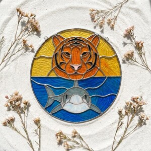 May include: A circular stained glass suncatcher featuring an orange tiger head set against a yellow background above a grey shark swimming in blue water. The piece is constructed with leaded glass segments and includes two small metal loops for hanging.