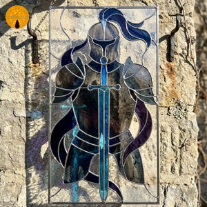 May include: Stained glass panel depicting a medieval knight in full armor, holding a sword. The artwork features shades of blue, purple, and gray, with a detailed design of the knight's helmet, armor, and flowing cape. The panel is framed with a metal border and hangs from chains.