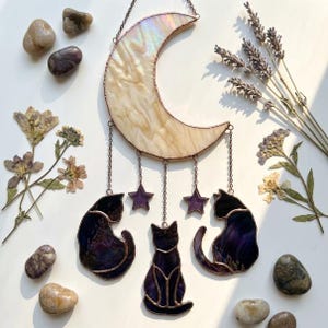 Op de afbeelding: Een hangende zonnevanger met een halve maan van iriserend wit glas, waaronder drie donkerpaarse glazen katten-silhouetten en twee kleine paarse glassterren hangen, verbonden door dunne metalen kettinkjes met koperkleurige soldeerlijnen.