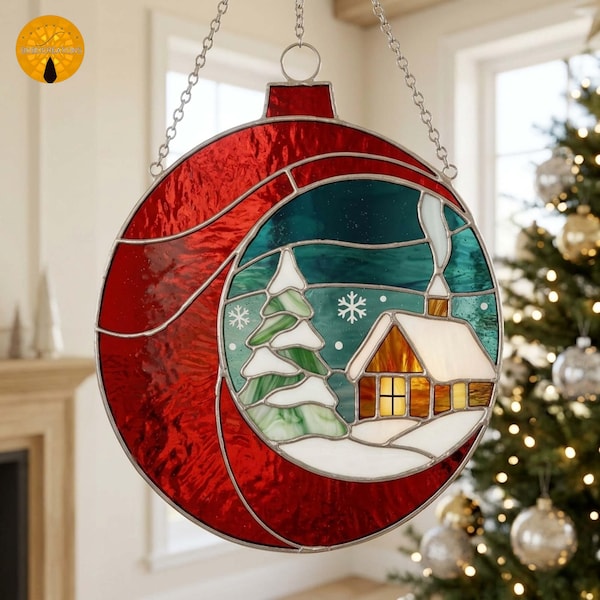 Puede incluir: Un adorno circular de vidriera con un borde rojo y una escena invernal central. La escena muestra una pequeña casa con ventanas amarillas brillantes, un tejado cubierto de nieve, un pino y copos de nieve blancos sobre un fondo verde azulado.