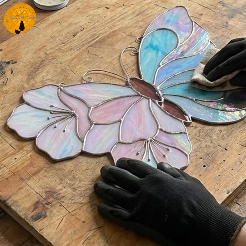 以下が含まれることがあります： 花柄デザインのステンドグラスの蝶。蝶はパステルピンク、ブルー、パープルの色合いで、茶色の体と銀色の金属の輪郭が特徴です。羽は花のような模様で詳細に描かれています。
