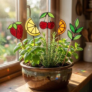 May include: A ceramic pot filled with herbs, adorned with decorative glass fruit and leaf garden stakes. The stakes feature a strawberry, lemon slice, cherries, orange slice, and green leaves. The pot sits on a wooden windowsill.