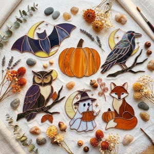 May include: A collection of stained glass ornaments featuring a bat, a pumpkin, a raven, an owl, a ghost wearing a witch hat, and a fox. The pieces are arranged on a light surface with dried flowers, small stones, and acorns scattered around.
