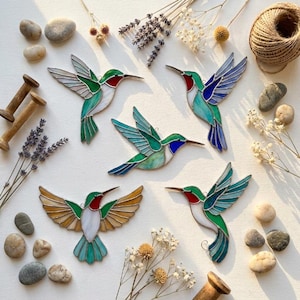 May include: Five stained glass hummingbird ornaments featuring various colour combinations of green, blue, white, and amber glass with dark metal soldering lines. Each bird is depicted in a different flight pose with wings spread, arranged alongside scattered stones, dried lavender, and twine.